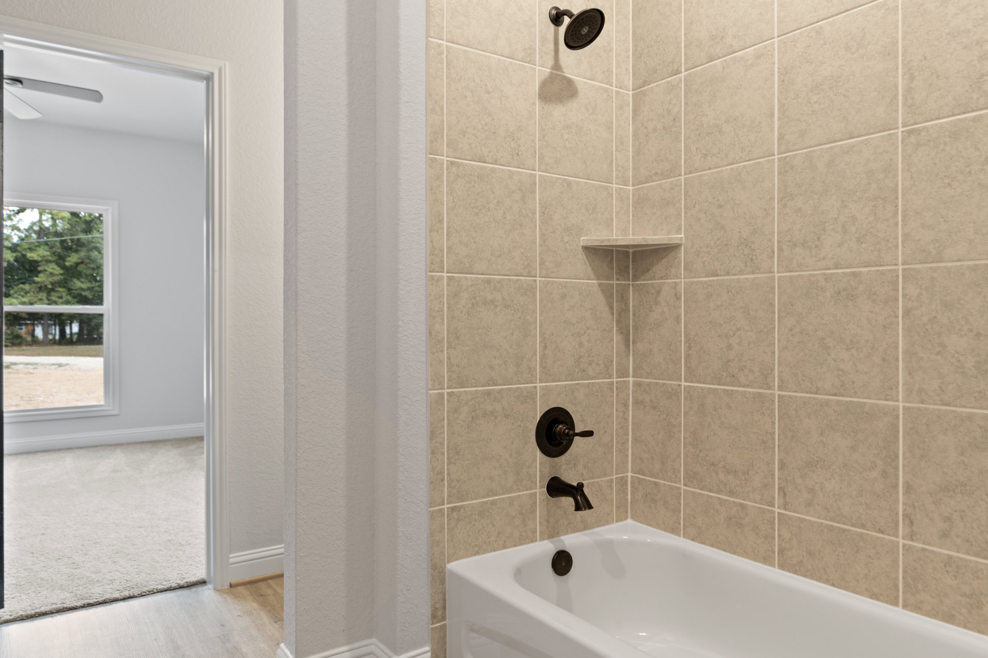 Freestanding white bathtub beside glass-enclosed shower with round showerhead, large window overlooking yard and trees, wood floor with white rug, chrome faucet and knob, light