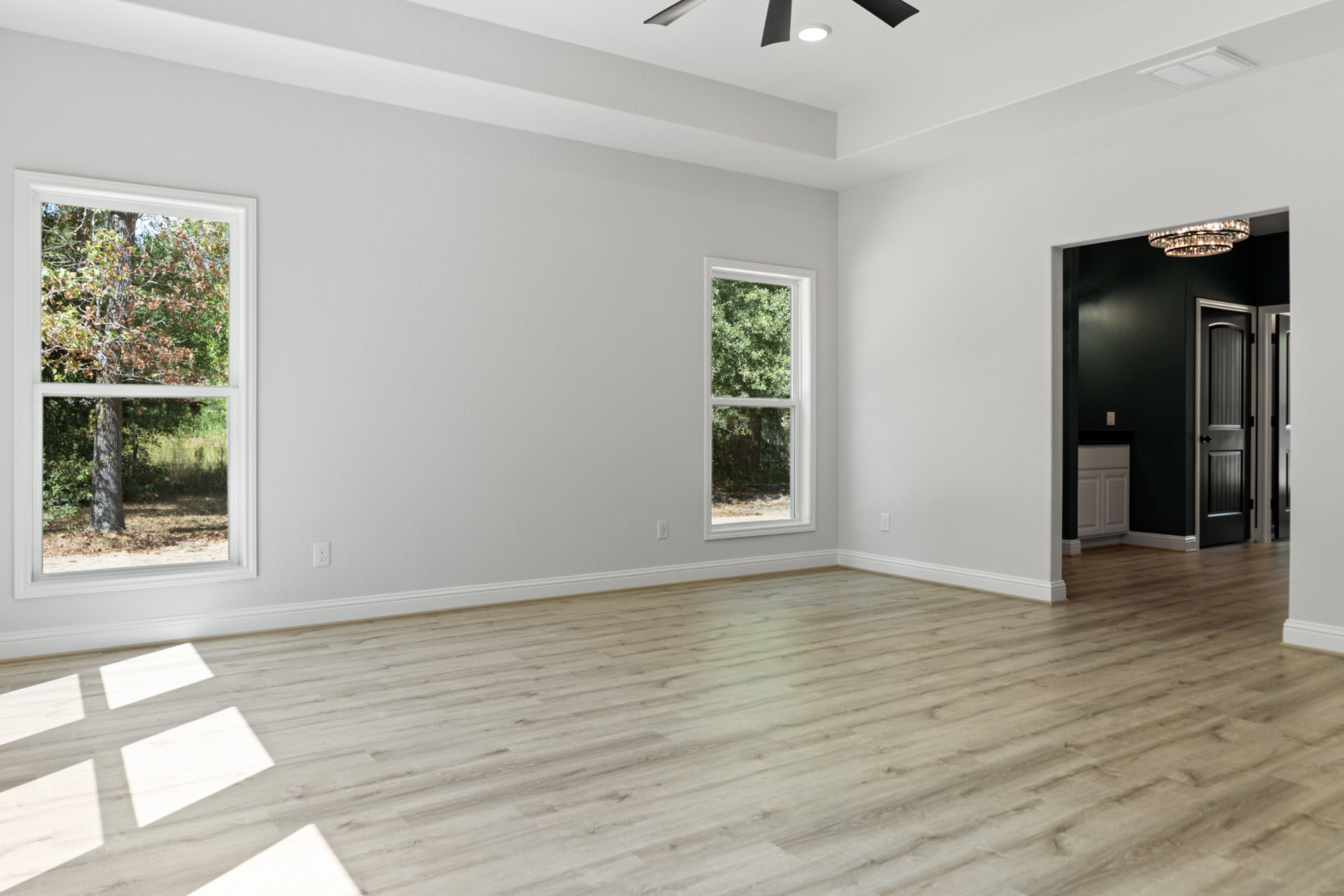 Wood flooring in a room with a ceiling fan, white cabinet, door, and large window overlooking trees and grass.