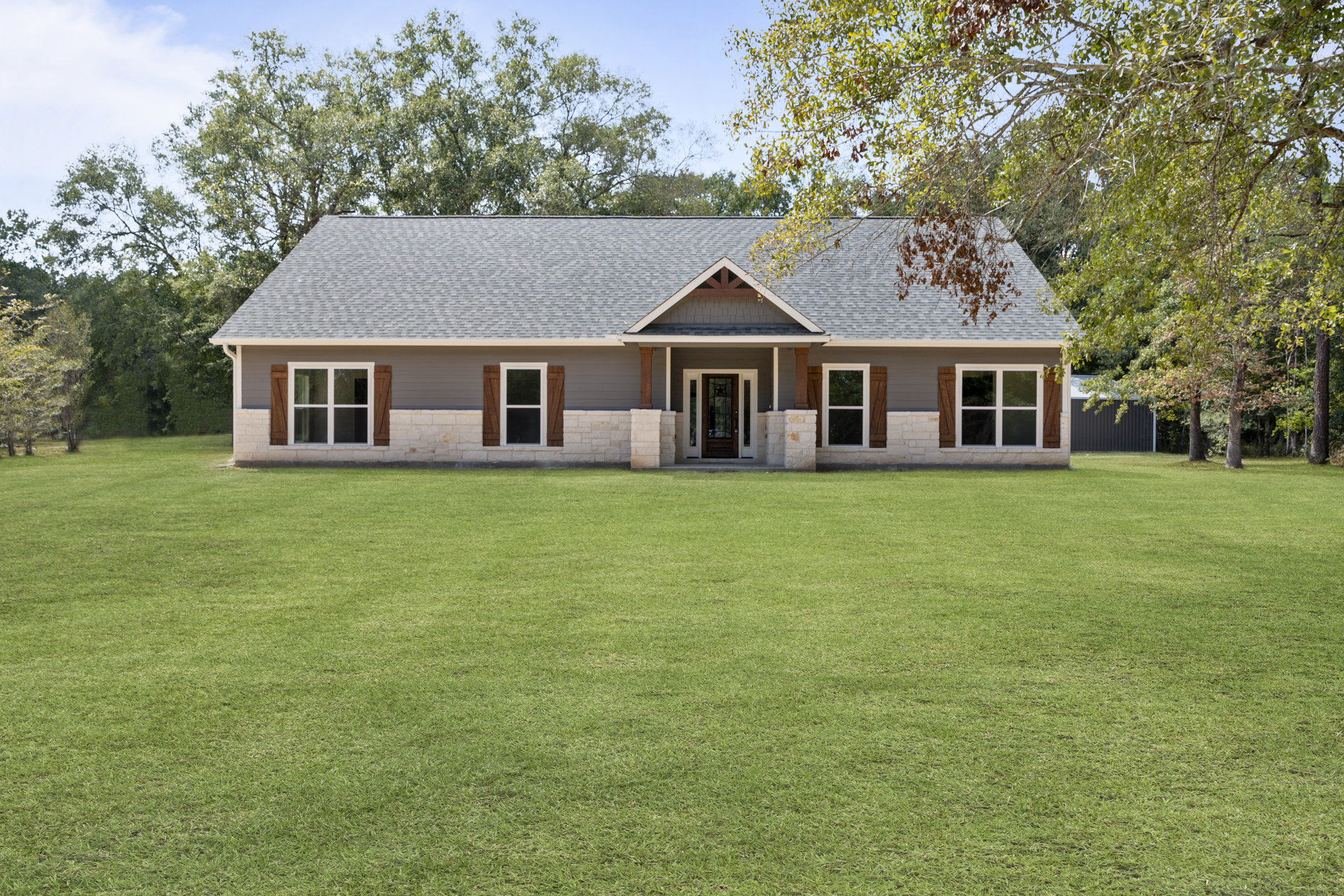 Stone exterior custom home with white-framed windows, glass door, and manicured green lawn bordered by trees and plants under a clear sky