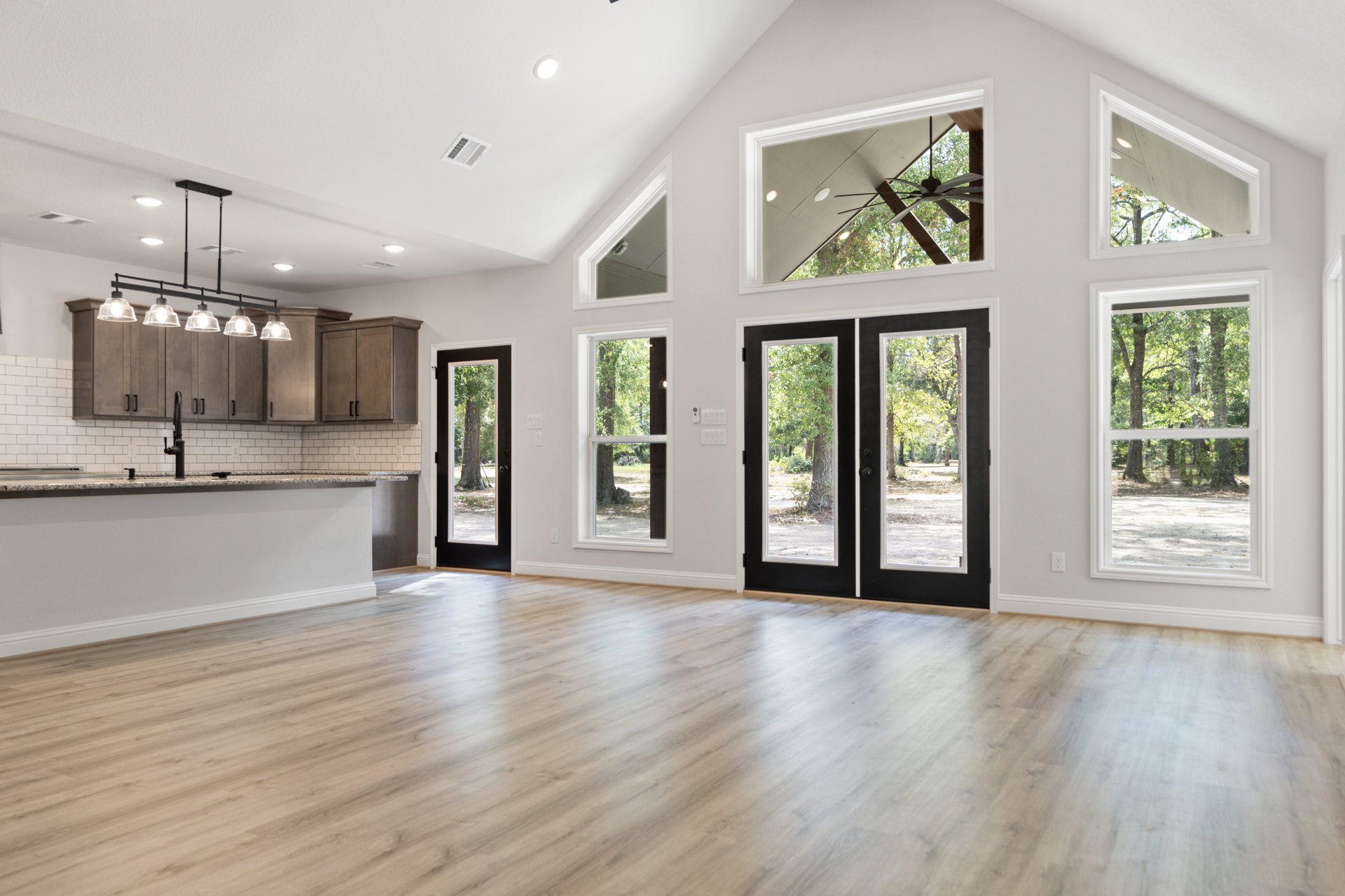 Open kitchen and living area with hardwood floors, white walls, ceiling fan, double glass doors, black interior door, and windows overlooking trees.