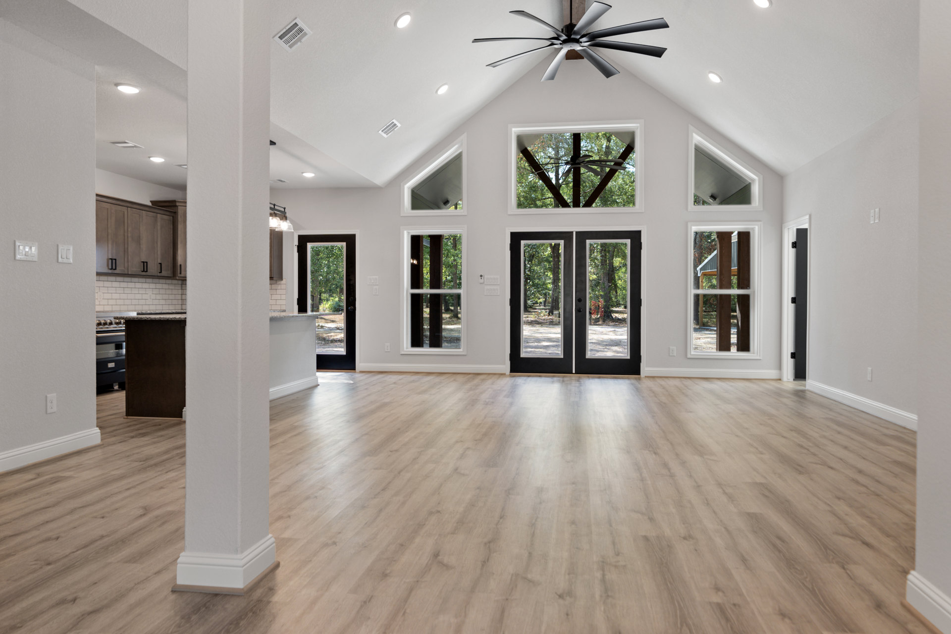 Wood flooring, black double doors with glass panes, ceiling fan mounted on wood post, large windows, white walls, built-in shelf