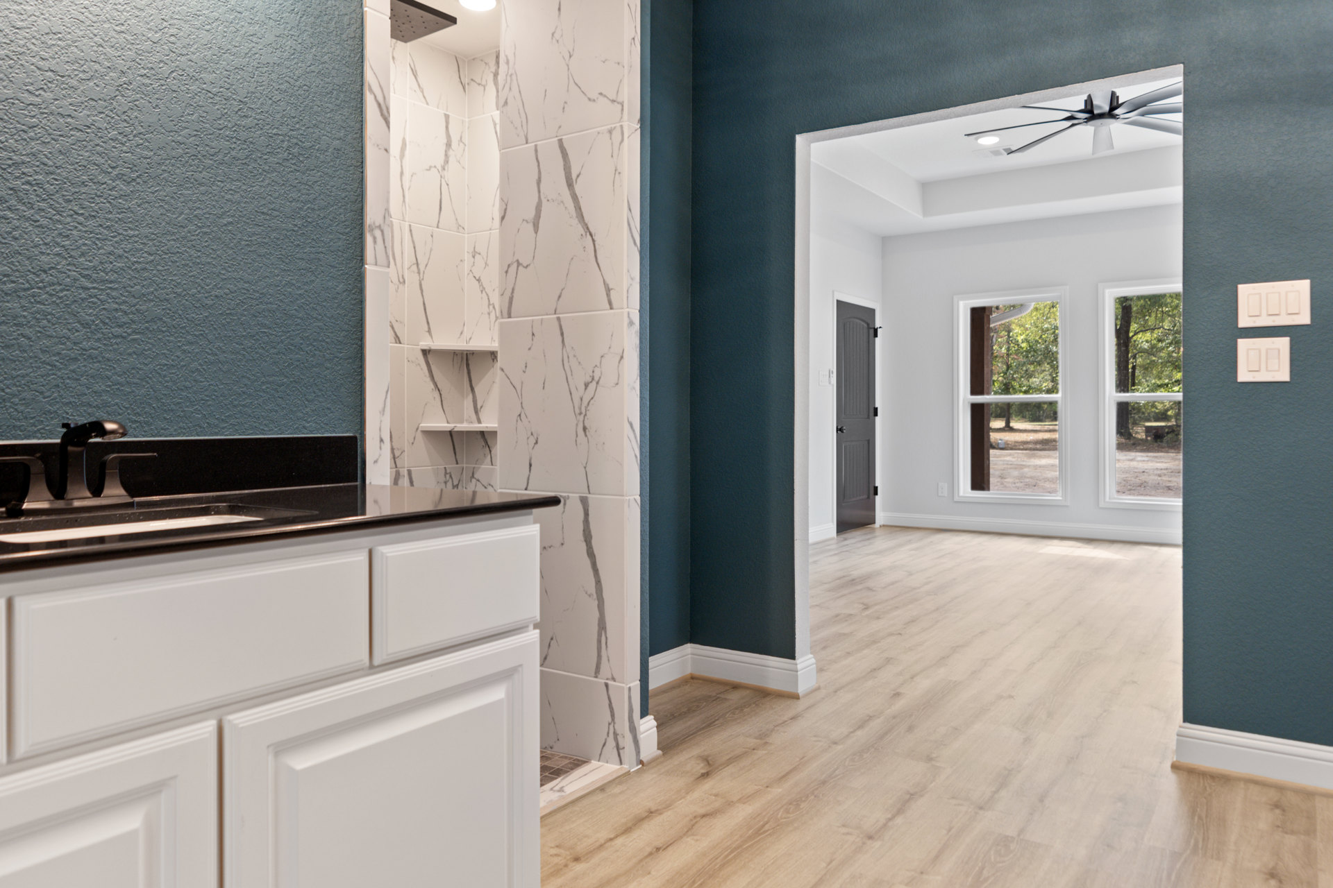 Marble accent wall, wood plank flooring, grey door with round doorknob, white cabinets and sink, ceiling fan, light switch on wall