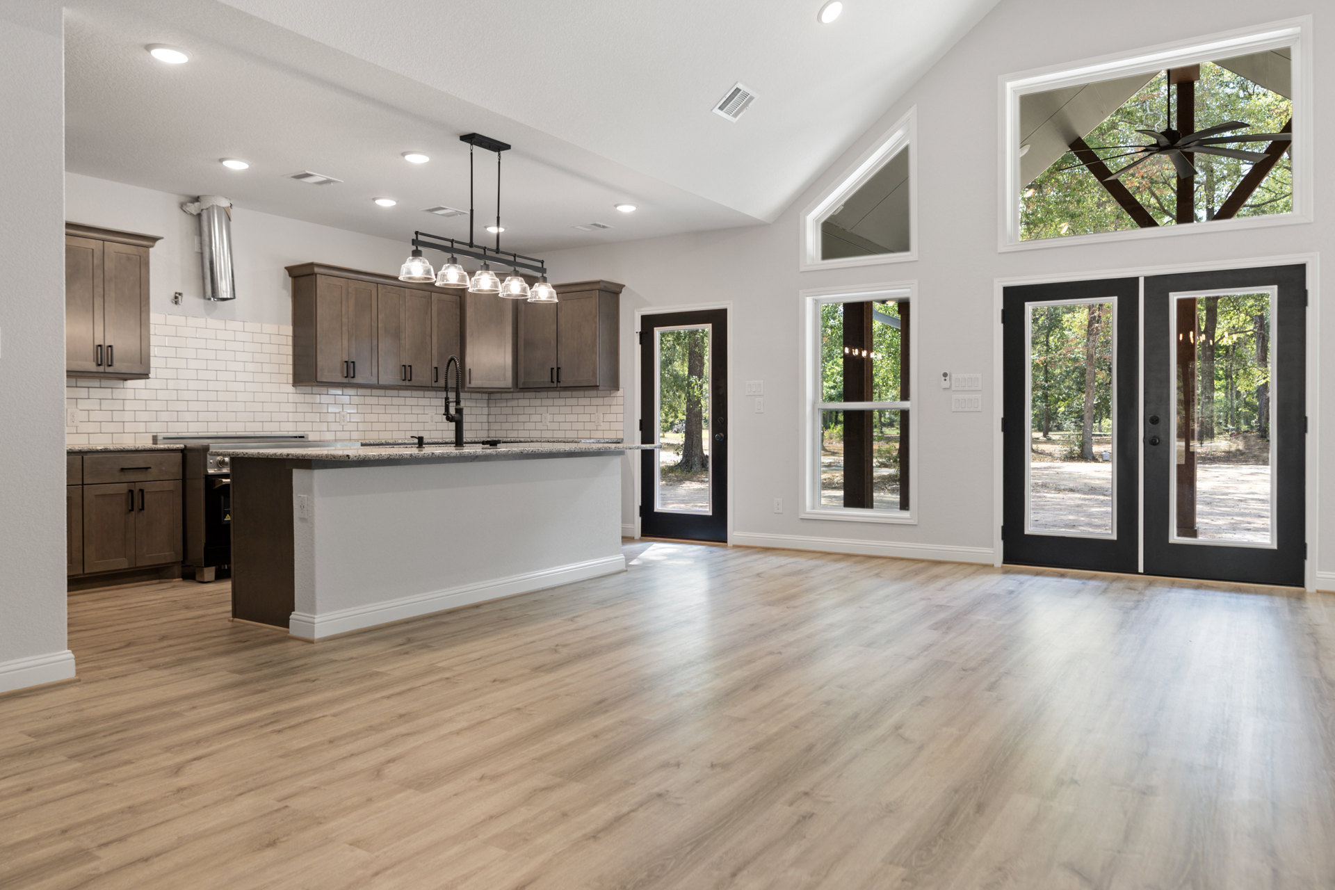 Open kitchen and living room featuring wood floors, white walls, marble countertop, black door, ceiling vent, cabinetry, and window overlooking trees