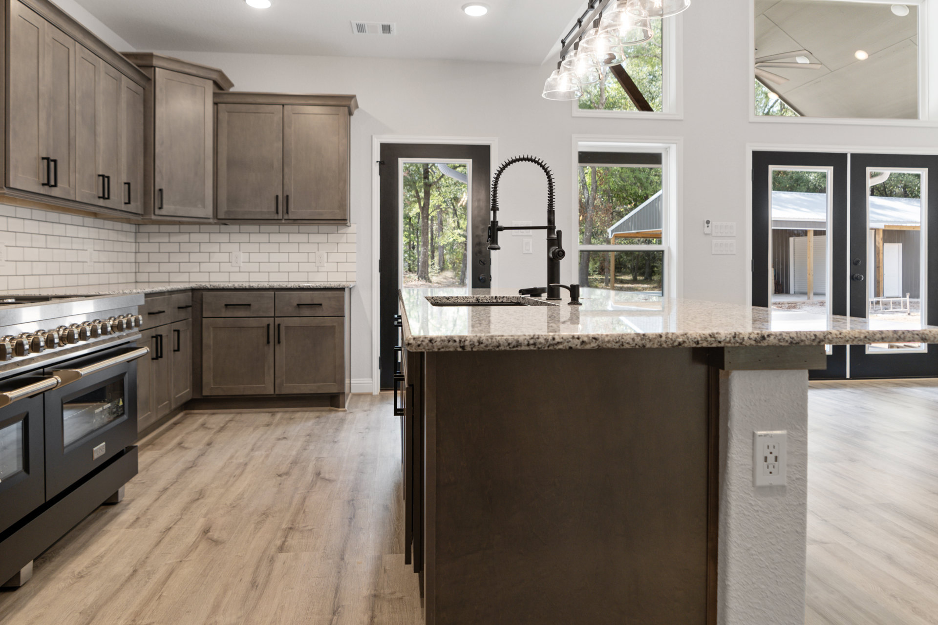 Open kitchen with wood flooring, white cabinetry, black oven, spring-style black faucet, quartz countertop, and a close-up of a white electrical outlet on the backsplash