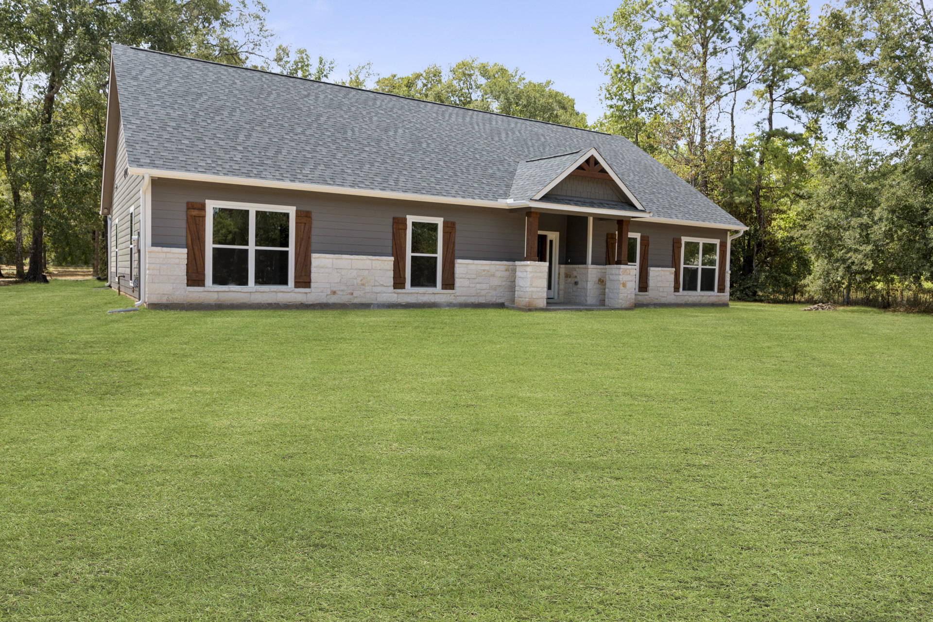 Stone-clad custom home with white-framed windows, expansive green lawn, mature trees, and clear sky