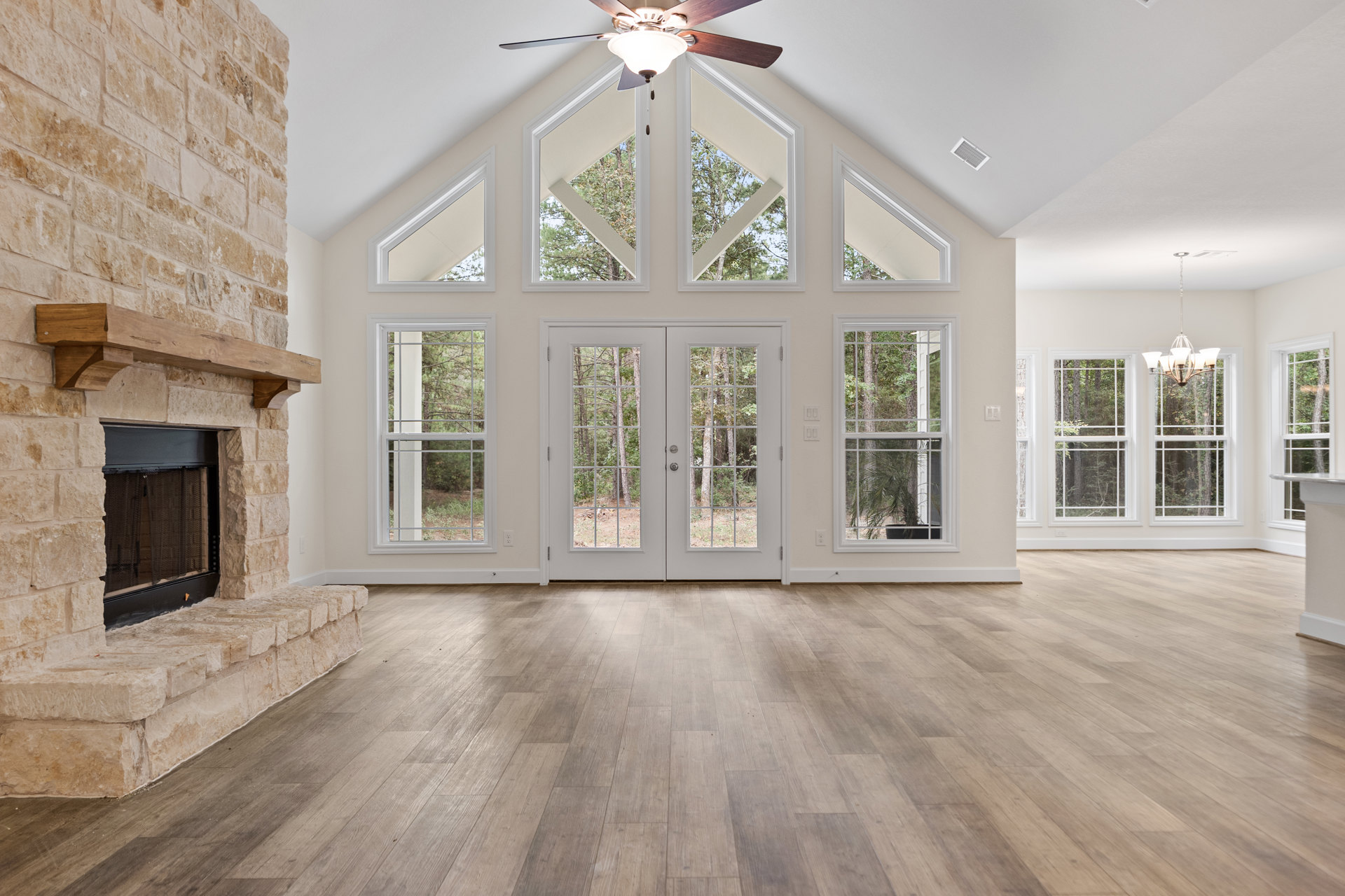 Wood floor and white walls in a living room featuring a brick fireplace with screen, ceiling fan with light fixture, double doors and window offering views of trees outside.