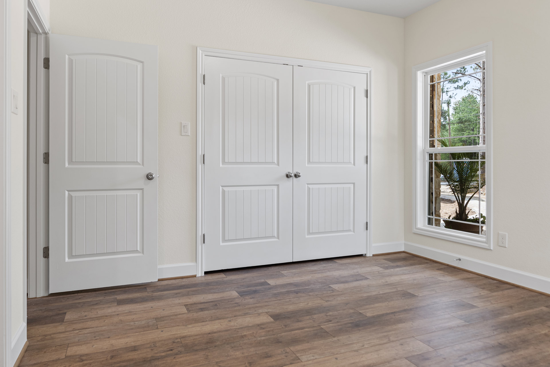 White double doors with silver knobs, single white door with silver knob, hardwood floor, potted palm tree, window showing trees and brick building