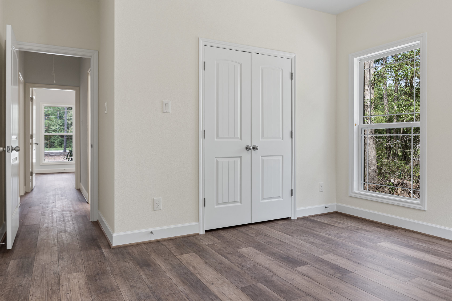 White double doors with silver knobs open to a room with wood flooring, white trim, and a window showing trees outside.