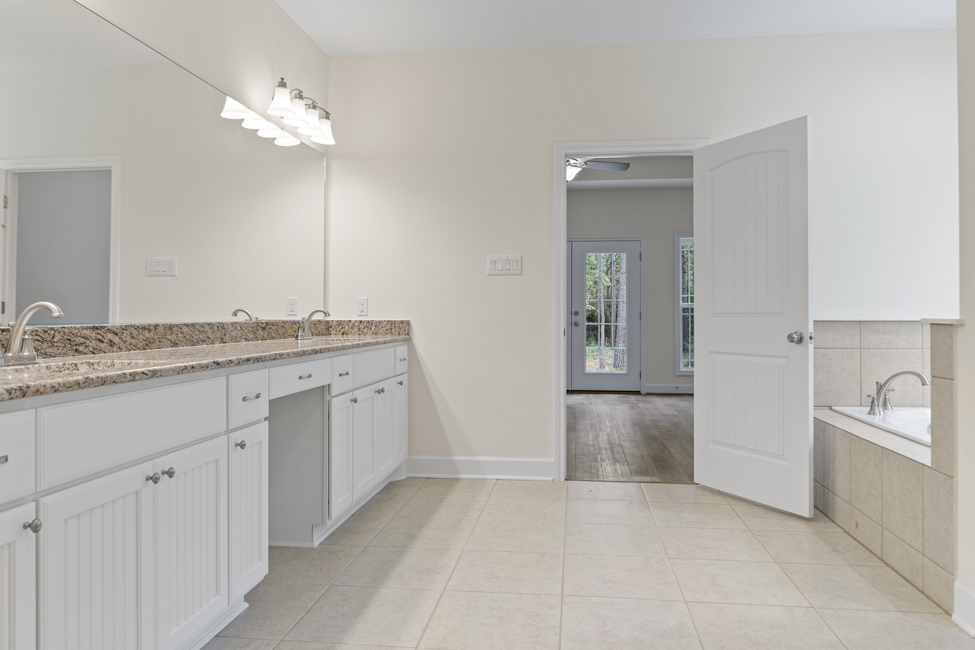 White bathroom cabinets with marble countertops, silver faucet, white tile floor, and a white door with a silver knob; door opens to a view of trees.