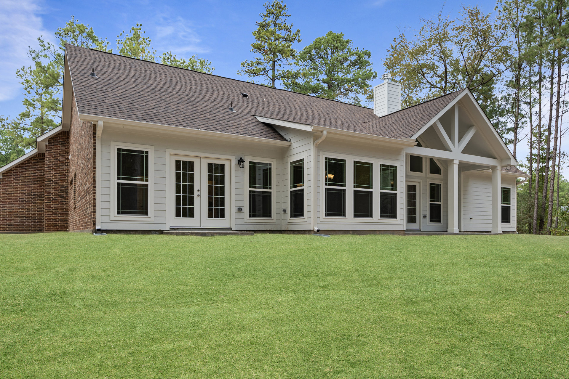 White two-story house with multiple windows, double glass-paneled doors, and a green grass lawn; security camera mounted near entry, trees and clouds in background.
