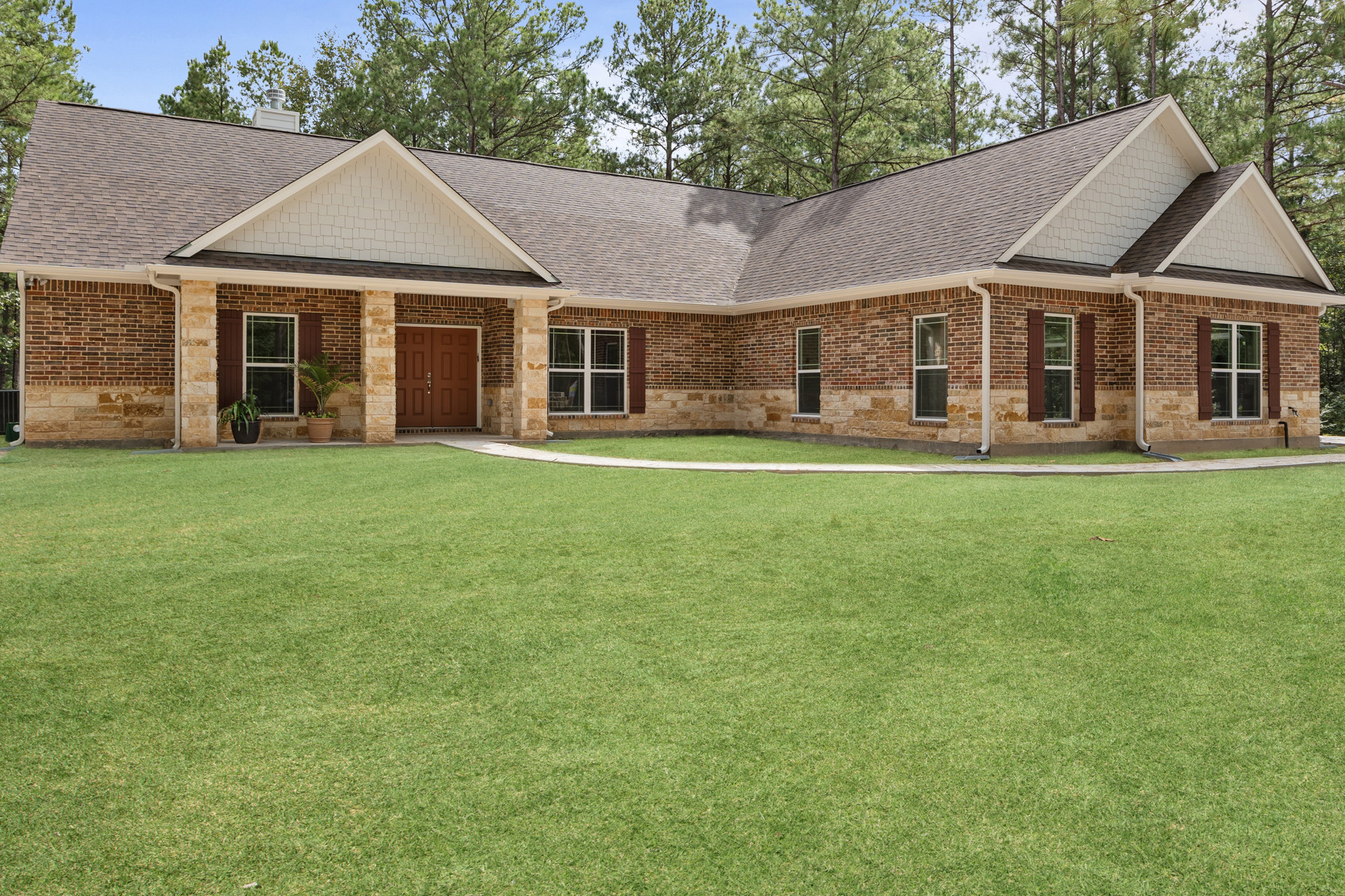 Brick house with double wooden doors framed by stone, glass-paned window, potted palm tree on porch, and manicured grass lawn.