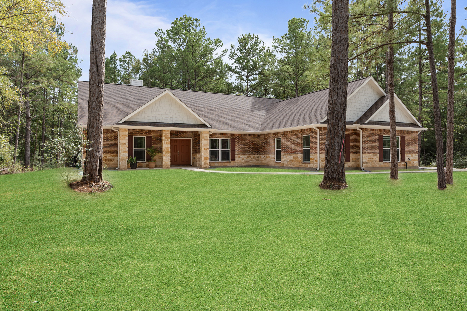 Brick house with white-framed windows, double front doors set in a stone wall, surrounded by green lawn and mature trees under a partly cloudy sky