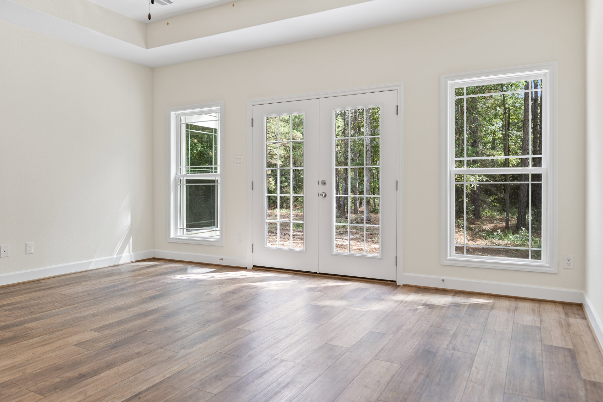 White-walled room with hardwood flooring, large windows framed in white, double doors opening to tree views, and daylight streaming through glass panes with white metal bars.