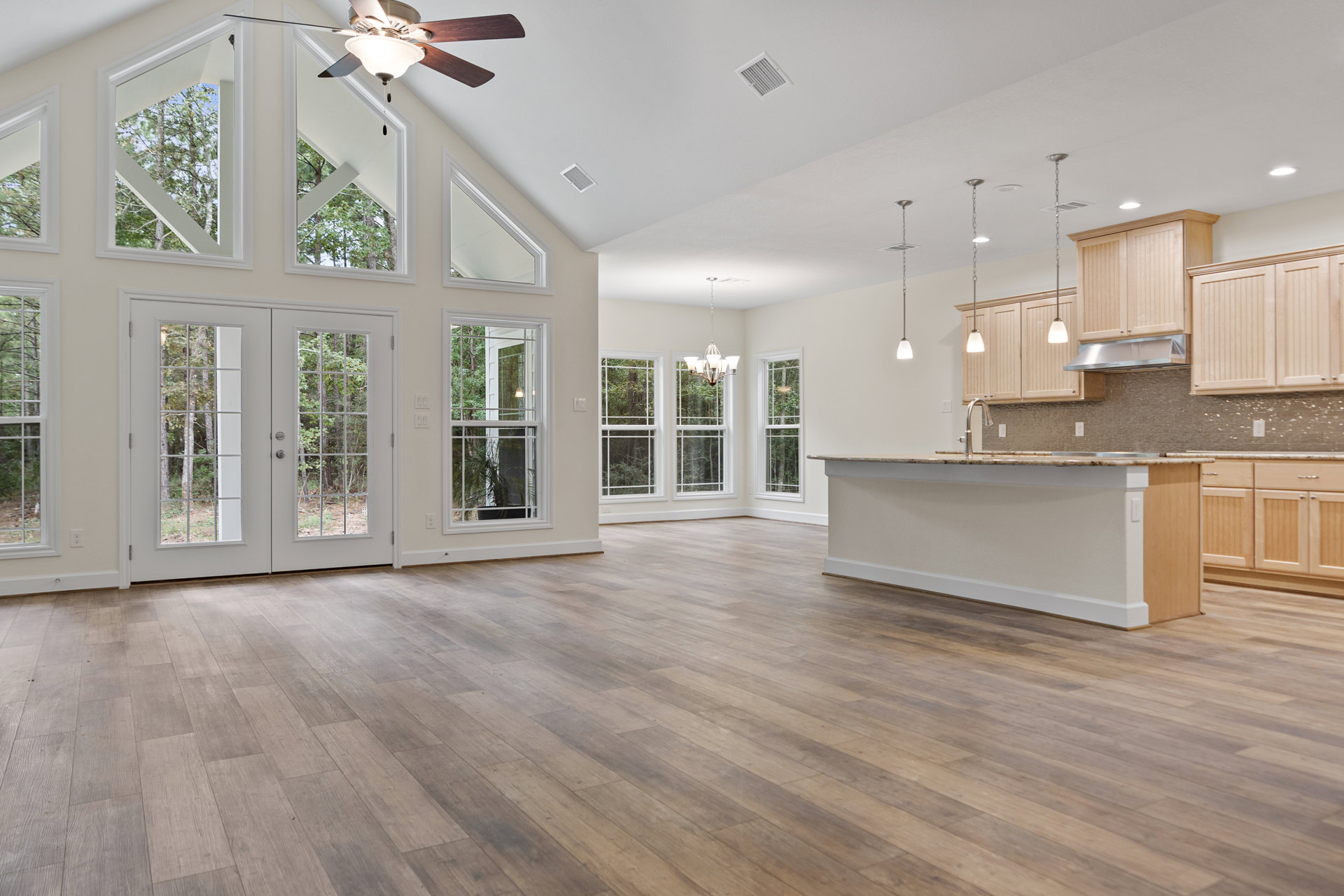 Open floor plan featuring wood flooring, white walls, white columns, kitchen cabinetry, dining area, ceiling fan with light fixture, ceiling vent, large window with white frame