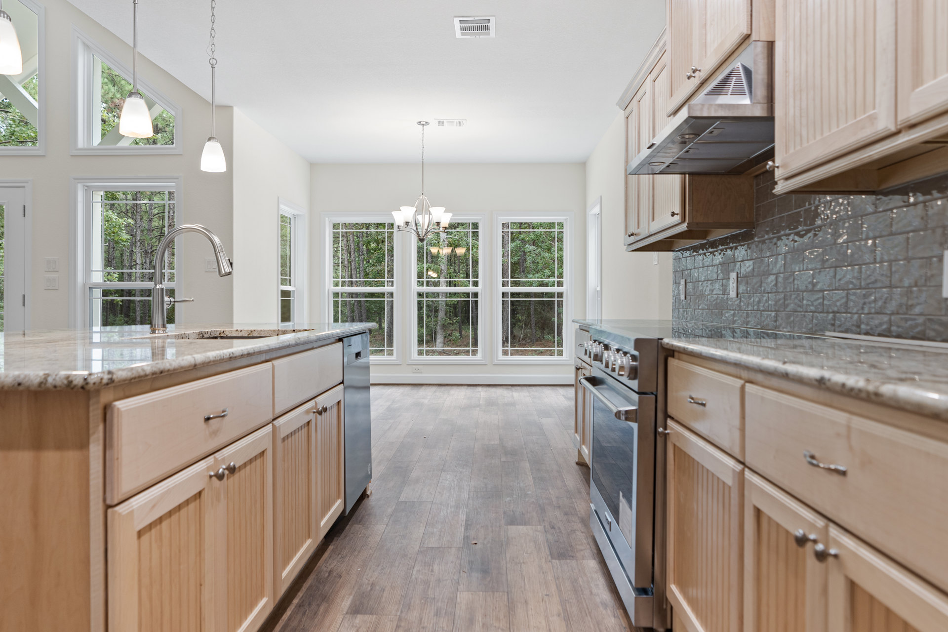 Kitchen with natural wood cabinets, matching wood floor, stainless steel appliances, stone countertops, modern chandelier, large window overlooking trees.