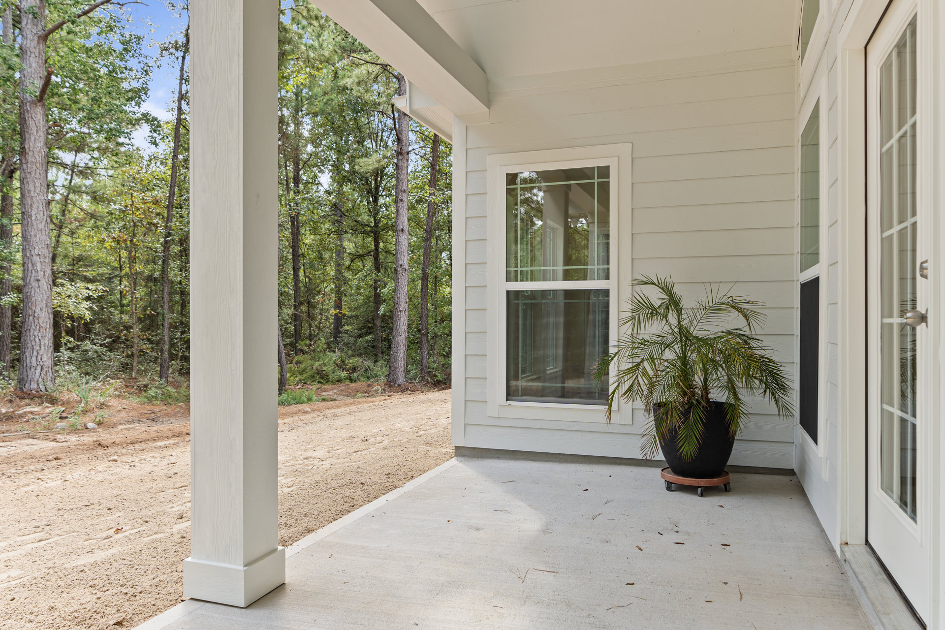 White siding house with glass pane windows, wooden porch featuring a potted plant, front door with paper tucked in handle, surrounded by trees and dirt road.