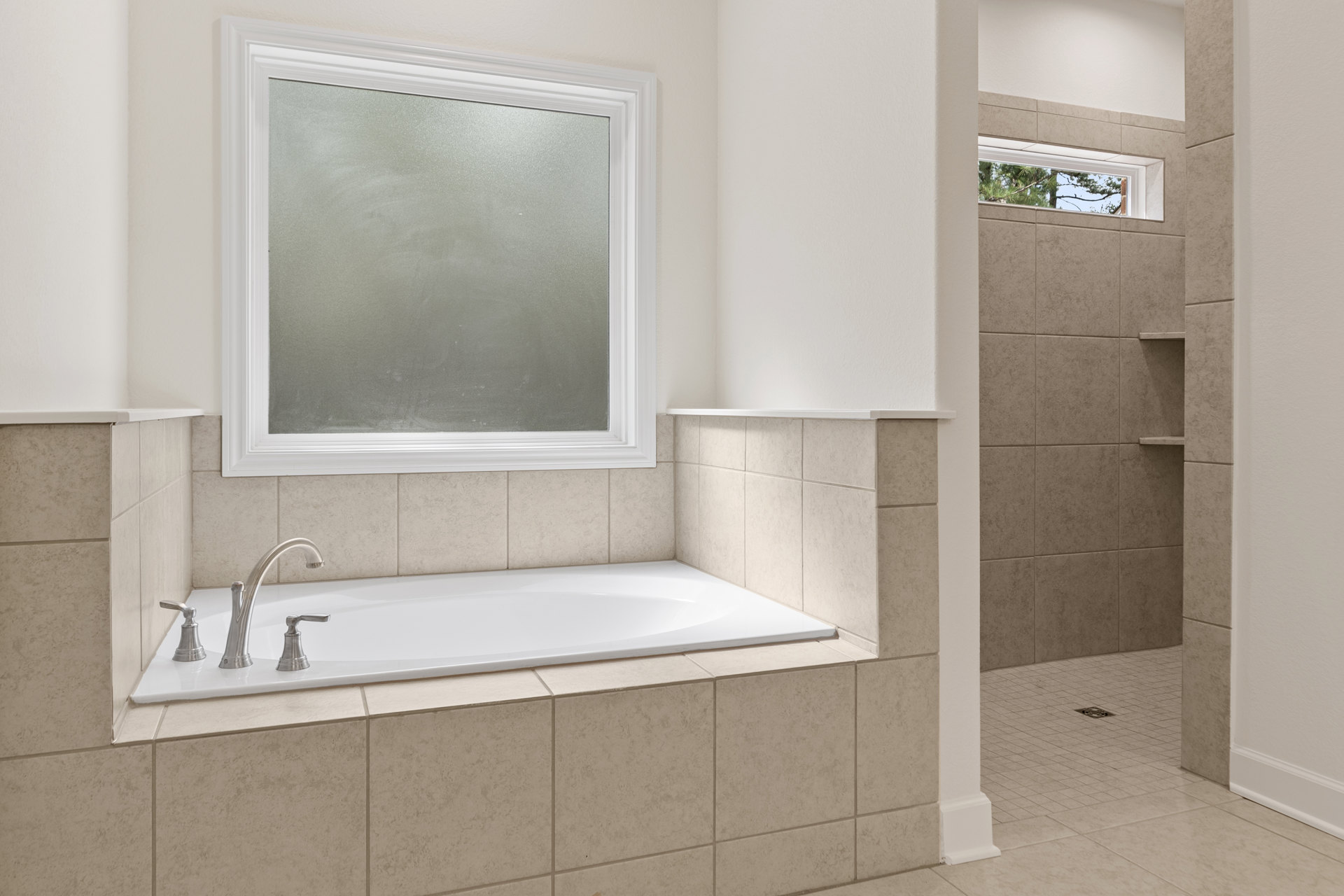 White bathtub with chrome faucet, tiled shower wall, frosted glass window, trees visible outside