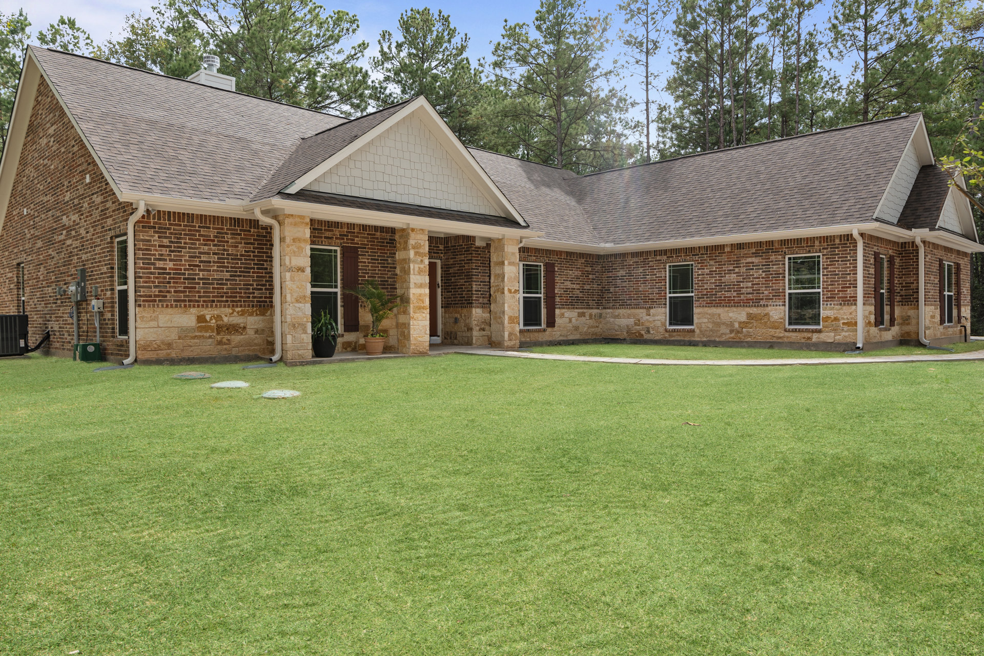 Red brick house with white-trimmed windows, potted palm tree near entry, green grass lawn, mature trees in background, roof vent visible above stone facade