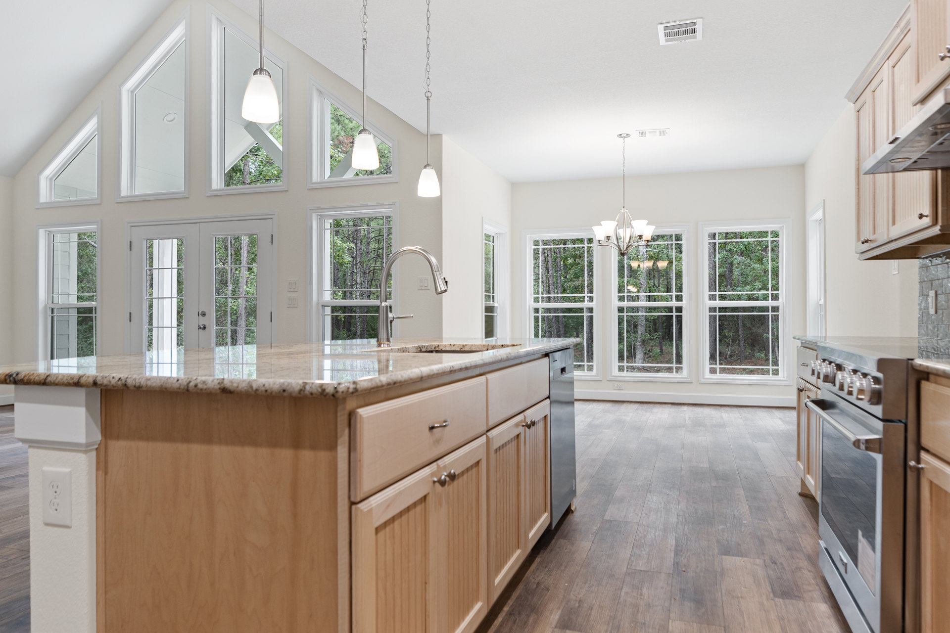 Spacious kitchen featuring a marble-topped island, large window with tree views, modern cabinetry, stainless steel oven, pendant lighting, and wide plank flooring