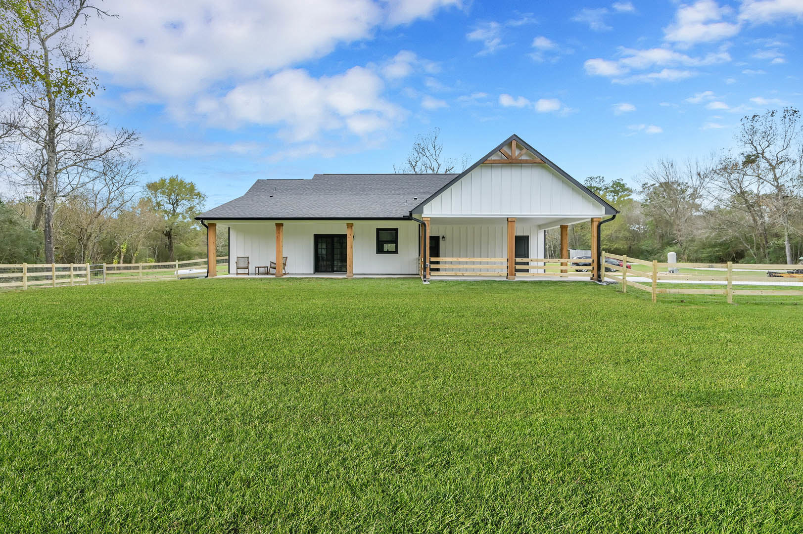 White house with black roof and black-framed windows, wooden fence enclosing green lawn, tree with bare branches, blue sky with scattered clouds