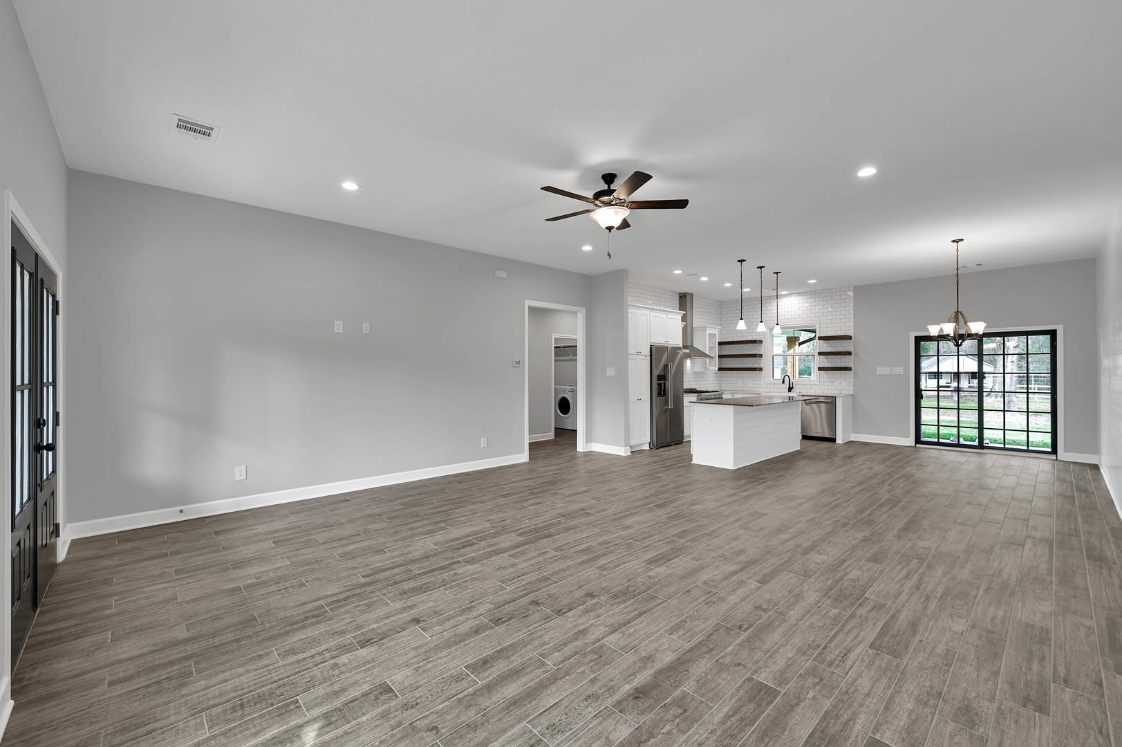Open-concept kitchen with white cabinetry, black countertops, wood laminate flooring, glass door, ceiling fan with light fixture, and plaster walls.
