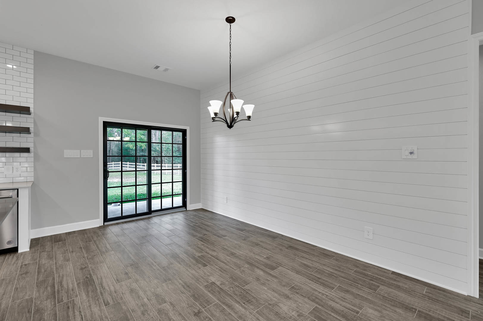 Wood-floored room with white walls, modern chandelier hanging from a chain, sliding glass door leading to fenced outdoor area, black-bordered white surface visible.