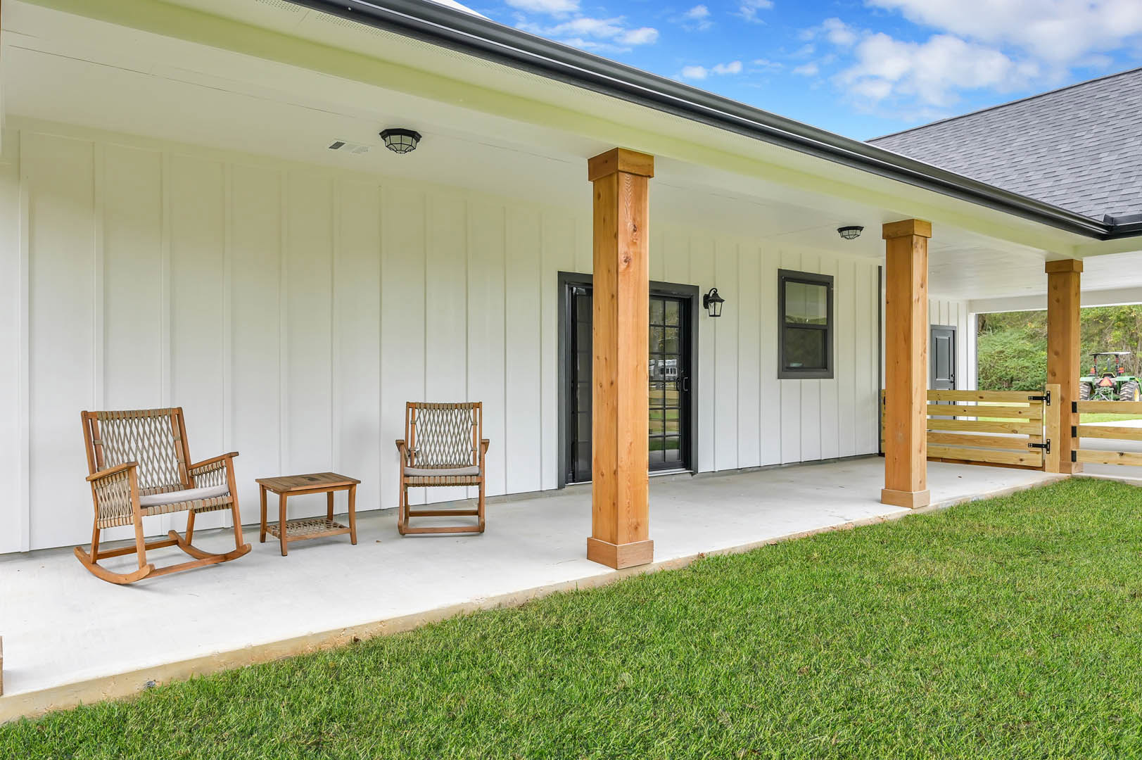 White exterior home with black-framed windows, wooden rocking chair and cushioned seat on porch, wooden table, green lawn, and shaded outdoor area.
