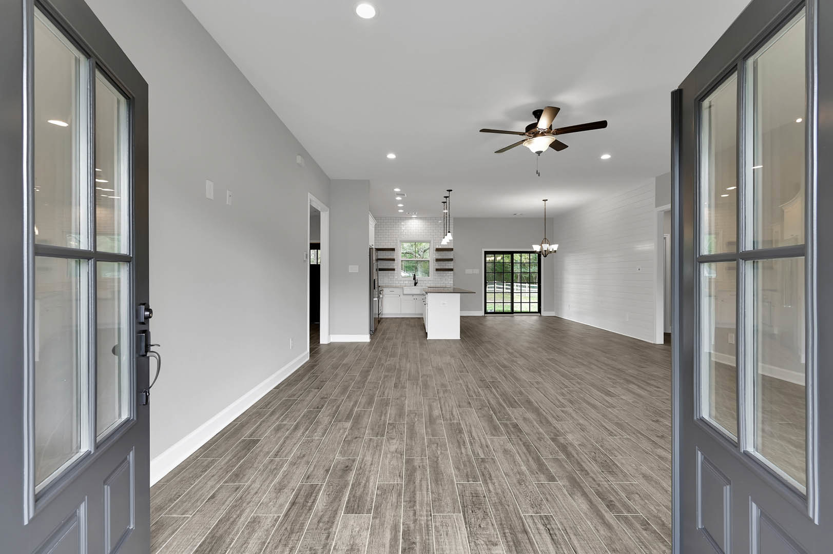 Wood flooring, white walls, ceiling fan with light fixture, sliding glass door opening to a fenced yard, large window providing outdoor view