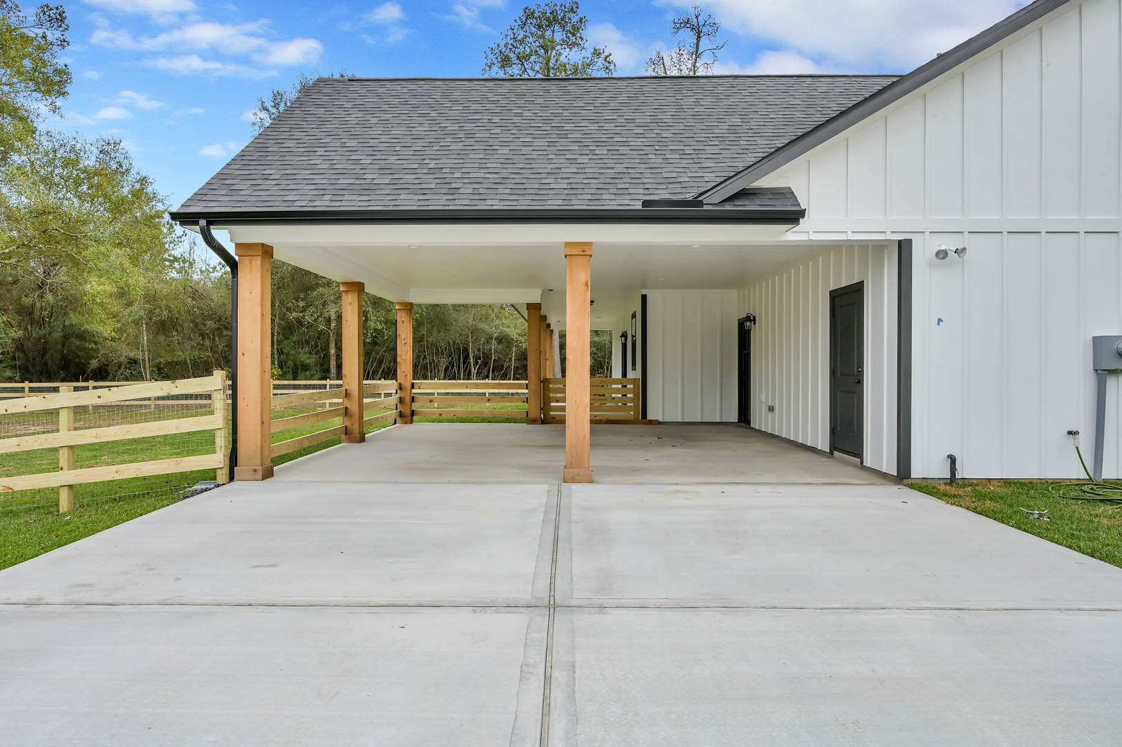 White siding home with covered patio, concrete driveway, wooden fence, and tree-lined roofline