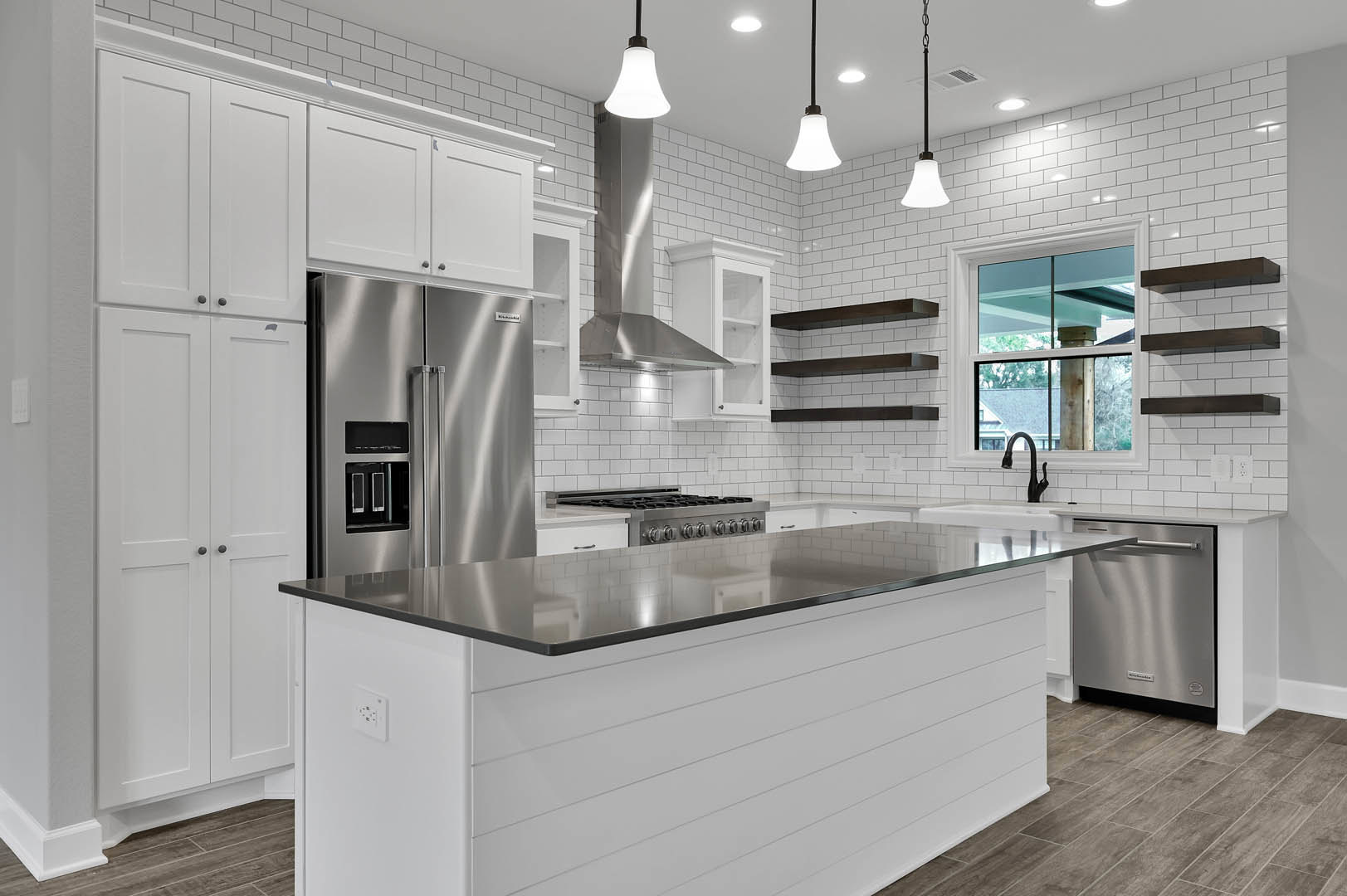 Spacious kitchen featuring a large central island with quartz countertop, white cabinetry, stainless steel refrigerator and dishwasher, glass-paned window above the sink, and light