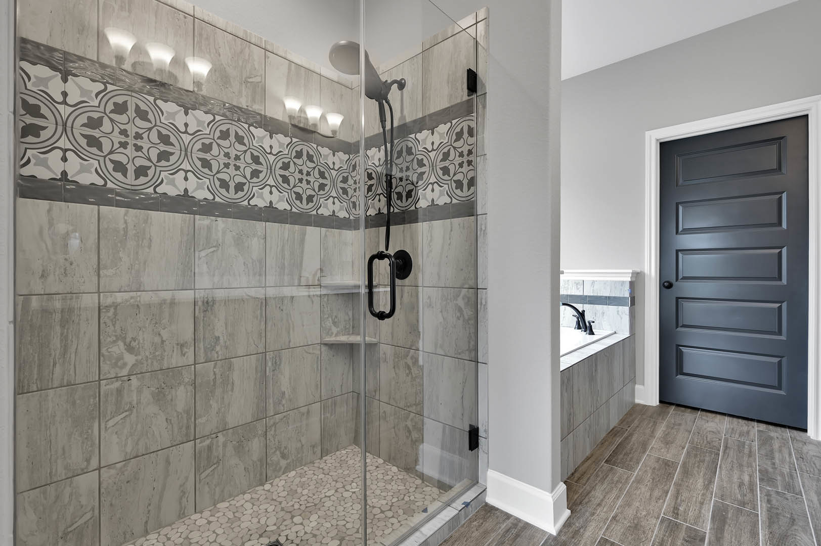 Walk-in shower with clear glass door, black metal handle, white tile walls, and light gray tile flooring