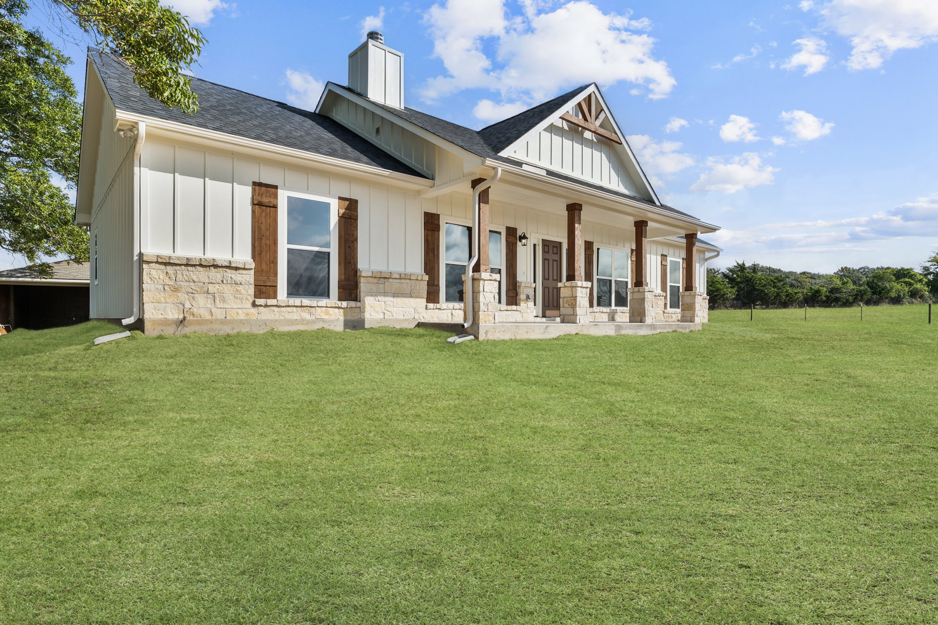 Stone house with white-framed windows, chimney, and stone wall, surrounded by green grass lawn and leafy tree under blue sky with clouds