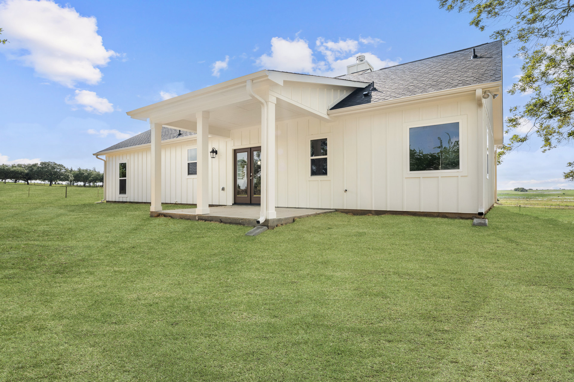 White siding house with covered porch, glass door reflecting trees, white framed windows, and green grass lawn under blue sky with clouds.