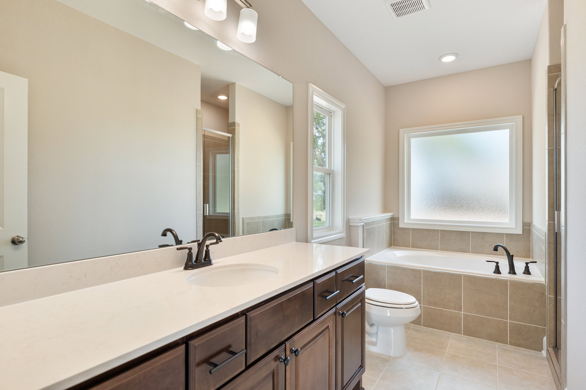 Bathroom with white porcelain sink and tub, closed white toilet, frosted glass window, wall-mounted mirror above sink, white lamp shade, tiled walls, and view of trees through