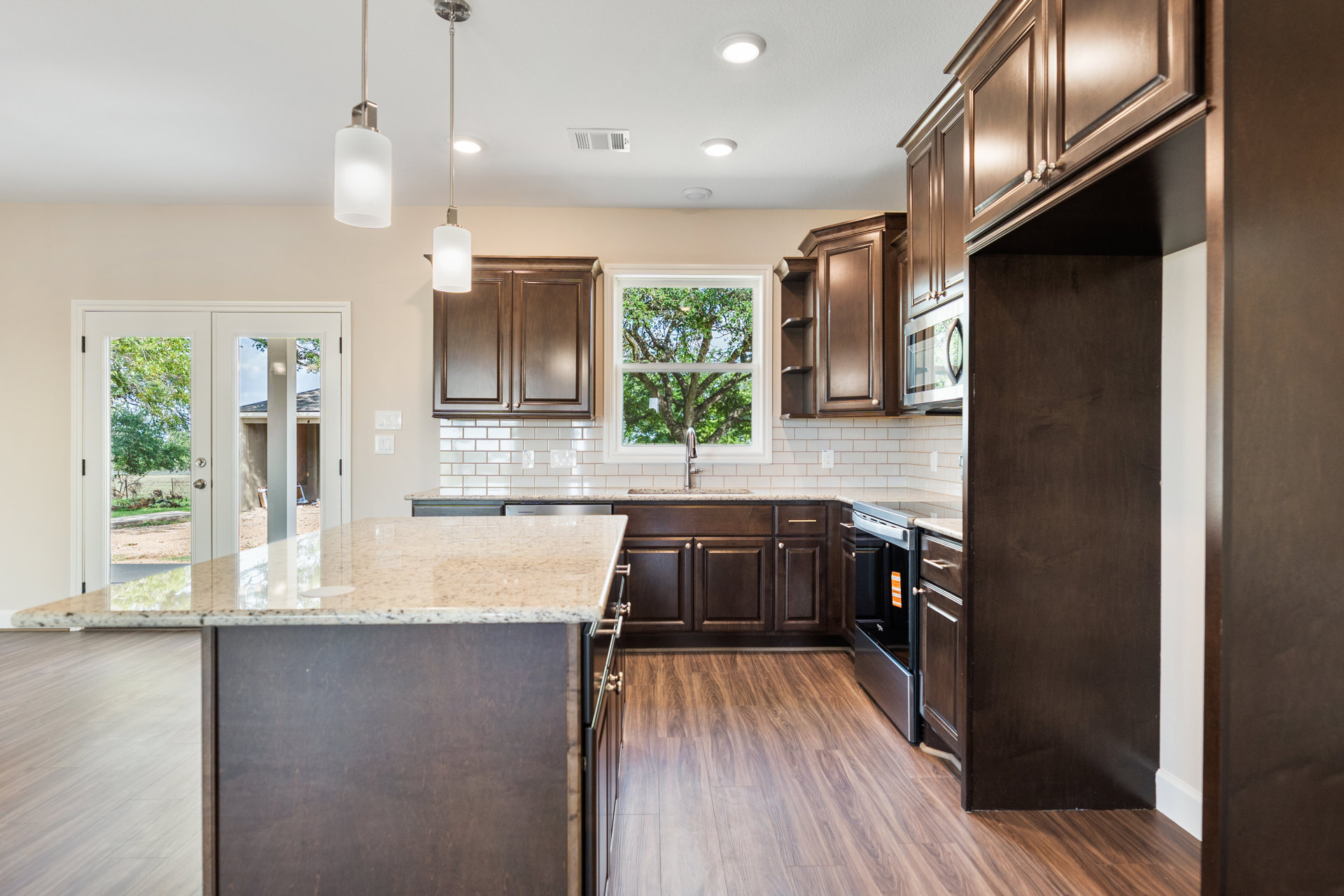 Dark wood cabinets and matching wood flooring in a kitchen with a marble-topped island, tree visible through window, modern pendant light fixture, white rectangular appliance, and