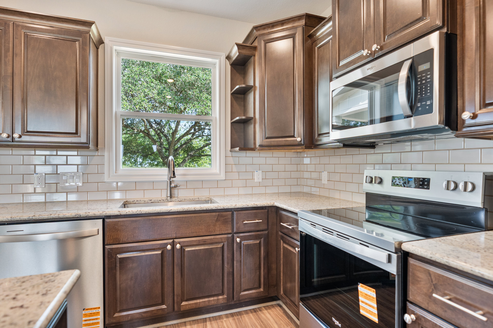 Brown wood cabinets, white tile floor, stainless steel stove and microwave, sink with chrome faucet beneath window showing leafy tree, light-colored countertops