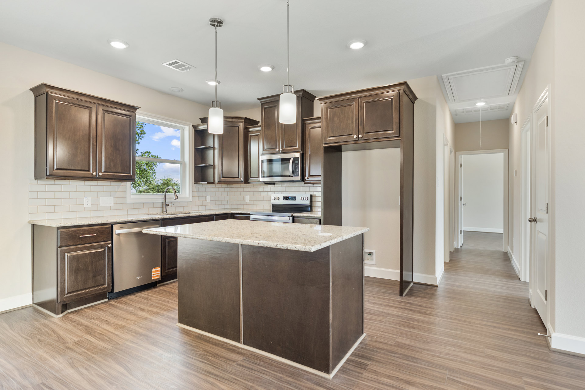 Spacious kitchen featuring a large marble-topped island, white cabinetry, stainless steel appliances including a microwave, pendant light fixtures, and wood flooring.