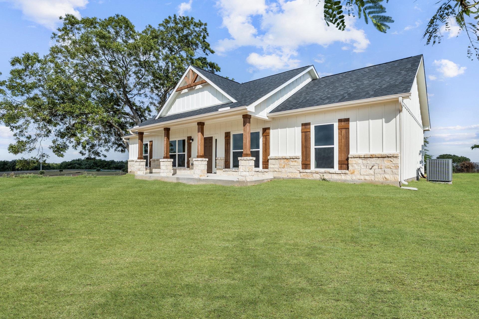 White house with stone porch and wall, large windows, green grass lawn, mature tree, cloudy sky in background