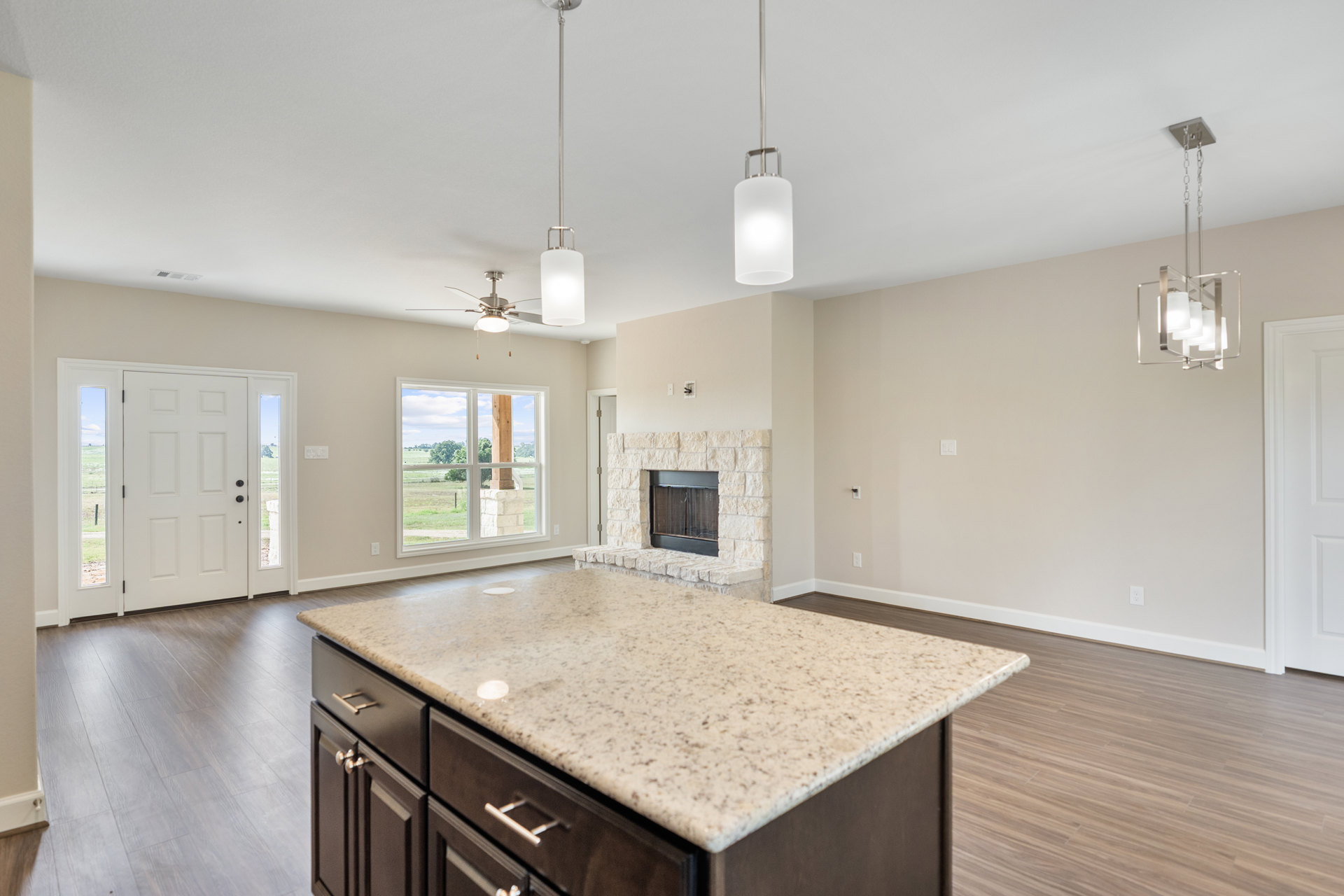 Quartz kitchen island with built-in sink, tile backsplash, white cabinetry, hardwood flooring, and large windows overlooking grassy field; fireplace visible in background.