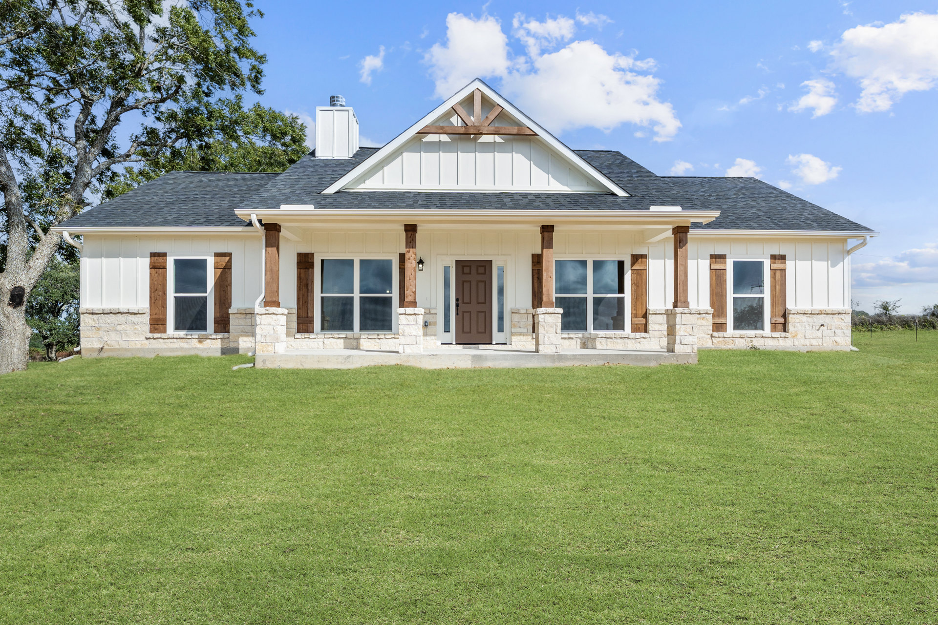 Stone exterior house with brown front door, white-framed windows, gray roof, and green lawn under partly cloudy sky