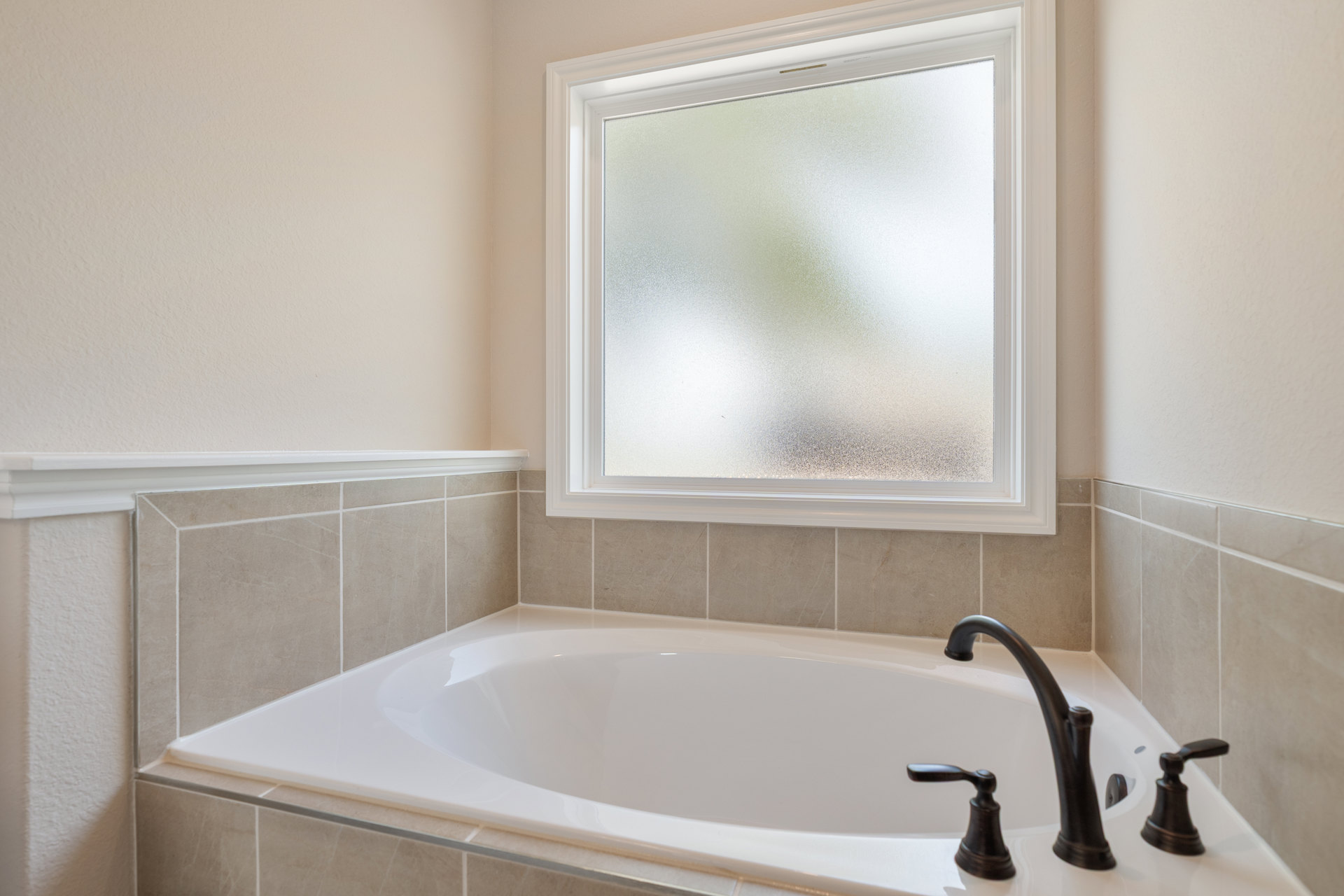 White freestanding bathtub beneath frosted glass window, surrounded by light gray wall tile, chrome faucet, and adjacent bathroom fixtures