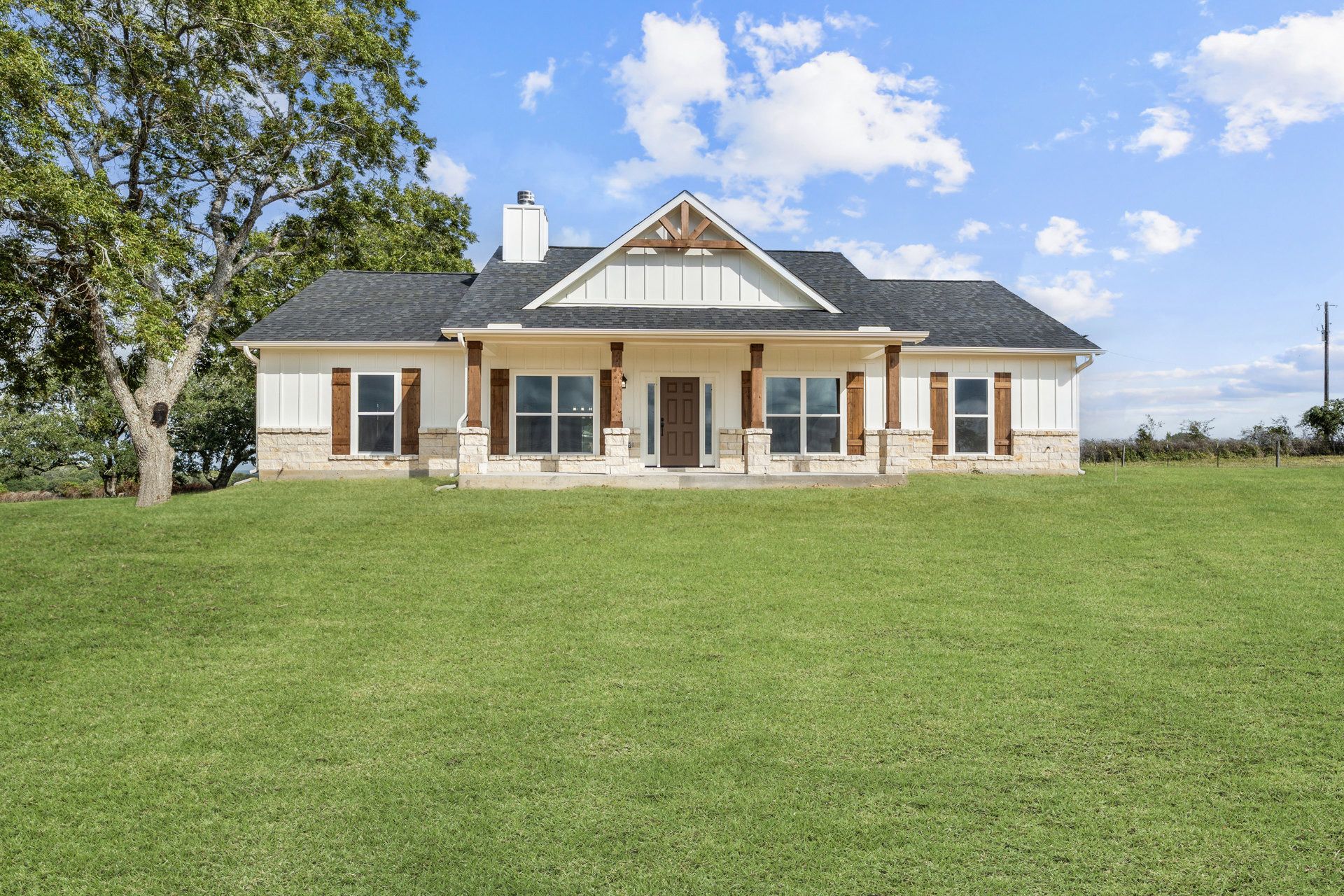 Stone-clad house with gabled roof and chimney, expansive green lawn, mature tree, large windows, brown front door, partly cloudy sky