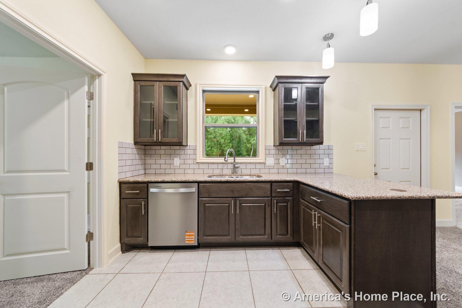 Dark wood cabinetry with glass-front upper cabinets, granite countertops, white subway tile backsplash, stainless steel dishwasher, single-handle faucet beneath a large window
