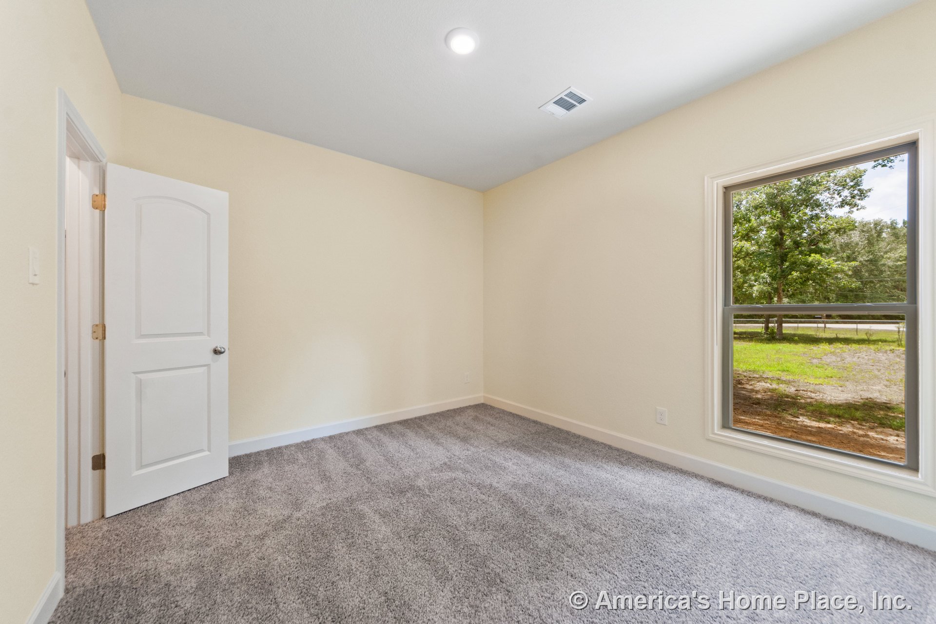 Bedroom with large window providing natural light, gray carpet flooring, cream-colored walls, white paneled door, recessed ceiling light, white baseboards, and wall vent.