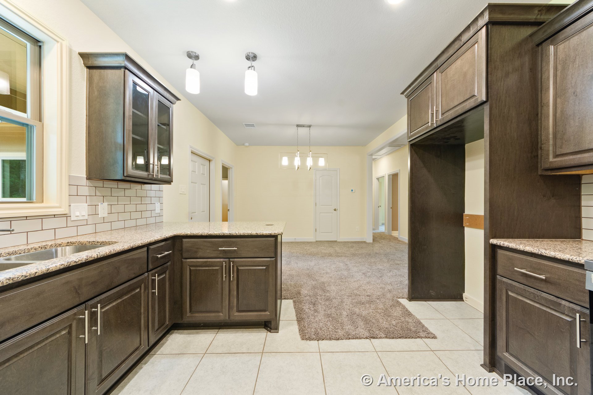 Dark wood kitchen cabinets with granite countertops, white subway tile backsplash, tile flooring, recessed and pendant lighting, window above sink, carpeted living area visible