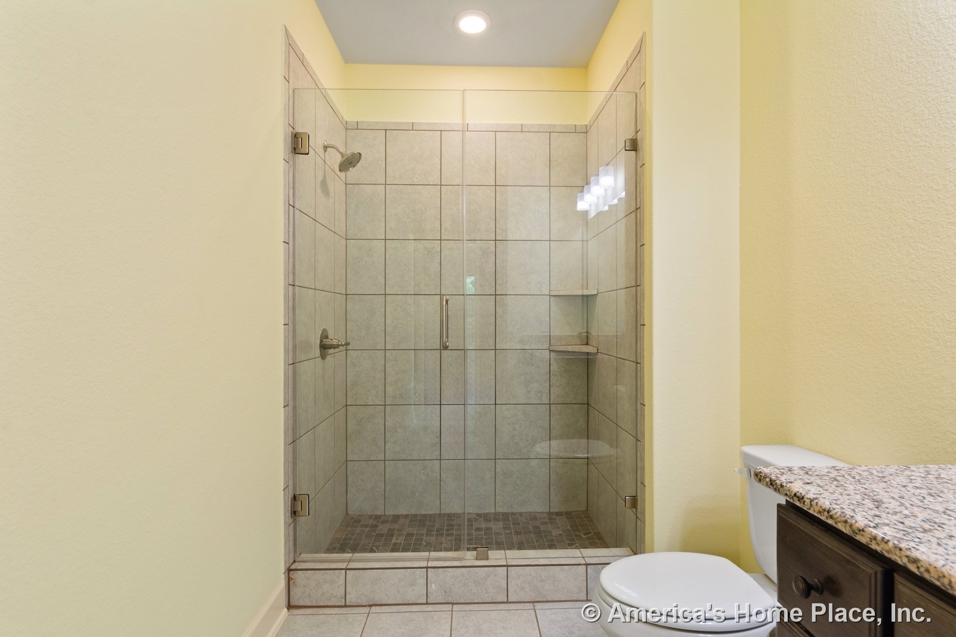Walk-in shower with glass enclosure, tile walls and floor, built-in shower shelf, granite countertop, recessed ceiling lighting, cabinetry, and toilet.