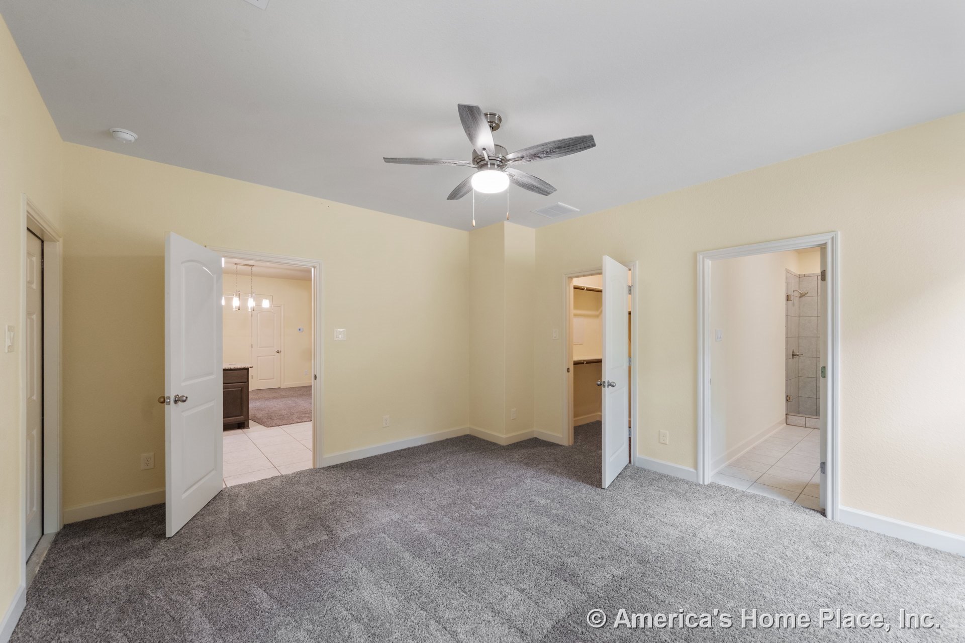 Gray carpeted bedroom with neutral walls, ceiling fan and light fixture, three white paneled doors leading to a walk-in closet, tiled bathroom, and hallway; baseboards and door
