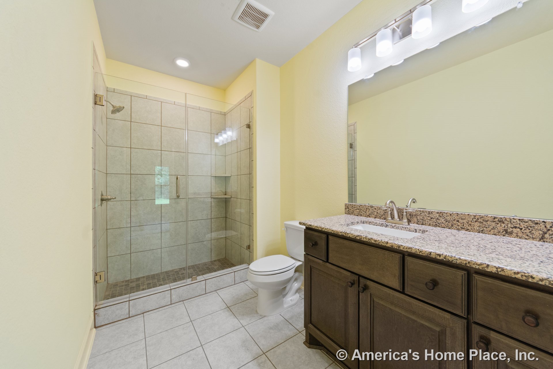 Walk-in tiled shower with glass enclosure, granite countertop vanity with dark wood cabinets, large wall mirror, tile flooring, built-in overhead lighting, and single sink adjacent