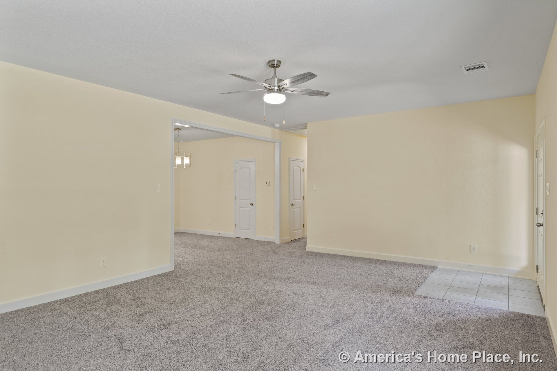 Carpeted living room with neutral walls, white trim and baseboards, ceiling fan with light fixture, open layout leading to a tiled entryway, and interior doors.