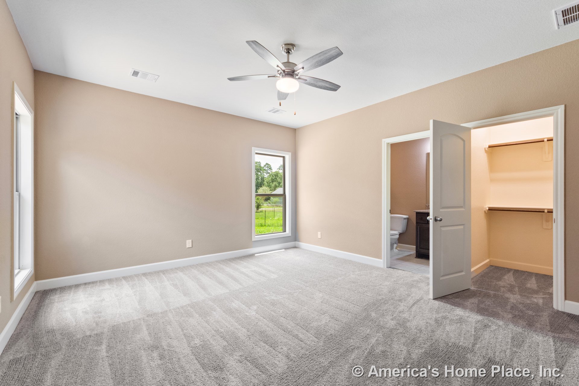 Bedroom with light gray carpet, beige walls, two large windows, ceiling fan and light fixture, en suite bathroom featuring a dark vanity, walk-in closet with built-in shelving
